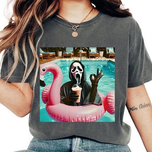 May include: Dark gray t-shirt featuring a graphic of a person in a Scream mask, holding a coffee drink while floating on a pink flamingo pool float in a swimming pool. The person is wearing black gloves and a black robe.