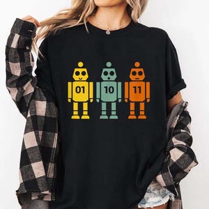 May include: Black t-shirt featuring three retro robot designs in yellow, teal, and orange. Each robot has a binary number on its chest: 01, 10, and 11. The robots have a simple, geometric design.