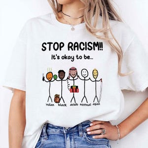 Op de afbeelding: Wit T-shirt met de tekst "STOP RACISM!! It's okay to be..." boven een afbeelding van een stokfiguur. De afbeelding toont stokfiguren van verschillende etniciteiten die elkaars handen vasthouden. De woorden "indian, black, arab, normal, asian" staan onder de figuren.