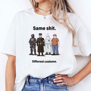 May include: White t-shirt with the text "Same shit. Different costume." above a cartoon illustration of four figures in different uniforms and attire. The figures include a person in tactical gear, a person in a military uniform, a person in a white robe, and a person in a casual outfit.