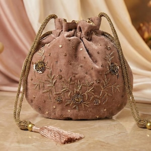 Op de afbeelding: Een stoffige roze fluwelen trekkoordtas met gouden geborduurde bloemmotieven en paillettenaccenten. De tas heeft gouden touwhandvatten en bijpassende kwastjes. Parfumflessen staan op de achtergrond.