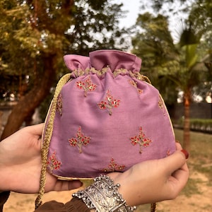 Può includere: Un sacchetto con coulisse color lavanda con finiture dorate e cordoncino dorato. La pochette è ricamata con motivi floreali rossi e dorati. La borsa è tenuta in mano.
