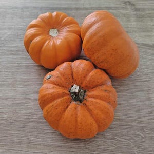 Puede incluir: Tres pequeñas calabazas naranjas con piel texturizada y tallos verdes. Las calabazas están dispuestas sobre una superficie de madera marrón claro. Las calabazas son de color naranja vibrante y los tallos son de color verde oscuro.