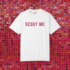May include: A white t-shirt with the text "SCOUT ME" in a burgundy color. The shirt has a crew neck and short sleeves. The background is a mosaic of red and pink tiles.