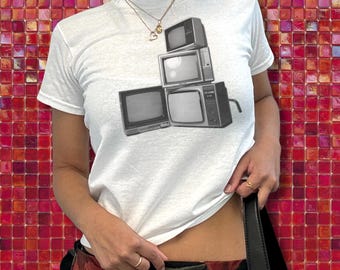 May include: White t-shirt featuring a black and white graphic of stacked vintage televisions. The shirt is a classic crew neck style. The background is a red and pink tiled wall.