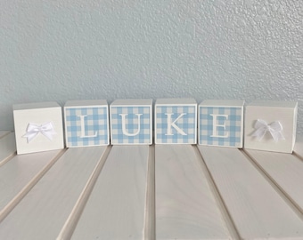 May include: Six square wooden blocks arranged in a row. The four centre blocks feature a light blue and white gingham pattern with the white capital letters L, U, K, and E. The two end blocks are solid white, each adorned with a small white bow.