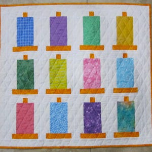 May include: A quilt with a white background and 12 colorful squares arranged in a grid pattern. Each square features a different patterned fabric, including blue, green, yellow, pink, and purple.
