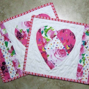 May include: Two pink and white quilted heart-shaped placemats with a floral pattern. The placemats have a pink and white striped border.