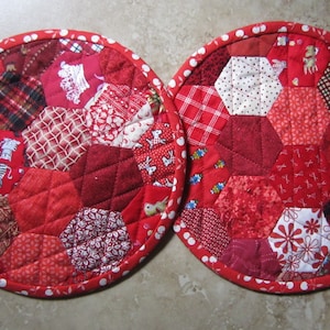 May include: Two red and white quilted pot holders with a hexagonal pattern. The pot holders are made of various red fabrics with white polka dots, plaid, and floral patterns.