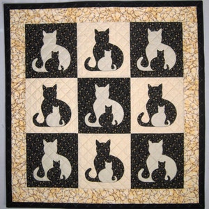 Op de afbeelding: Een quilt met een zwart-wit geruit patroon met negen silhouetten van katten. Elke kat houdt een kleinere kat in zijn armen. De achtergrond is crèmekleurig met een zwart bloemenpatroon.