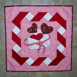 May include: A pink and red quilted wall hanging with a white envelope and two hearts. The envelope has a red ribbon and the hearts are brown with pink floral designs.