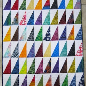 May include: A colorful quilt featuring a repeating pattern of triangles in various colors and patterns, set against a white background. The triangles include shades of red, orange, yellow, green, blue, purple, and black, creating a vibrant and eye-catching design. The quilt is bordered with a dark edge.