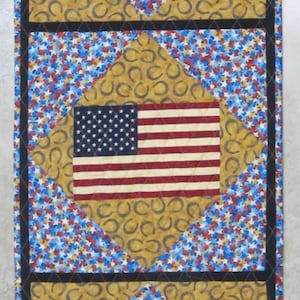 May include: A quilted table runner featuring three American flags. Each flag is set within a diamond shape, surrounded by blue and white stars and a gold horseshoe pattern. The runner is bordered with a black trim.