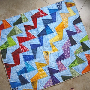 Może przedstawiać: Ręcznie robiona kołdra z jasnoniebieskim tłem i zygzakowatym wzorem z kolorowych trójkątów. Trójkąty są w odcieniach czerwieni, pomarańczu, żółci, zieleni, niebieskiego i fioletu, z różnymi wzorzystymi tkaninami. Kołdra jest obramowana paskowanym materiałem.