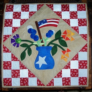 Puede incluir: Un bloque de colcha patriótico con un fondo a cuadros rojo, blanco y azul. Un jarrón azul con una estrella blanca se encuentra en el centro, lleno de flores azules y verdes. Una pequeña bandera estadounidense se muestra sobre el jarrón.
