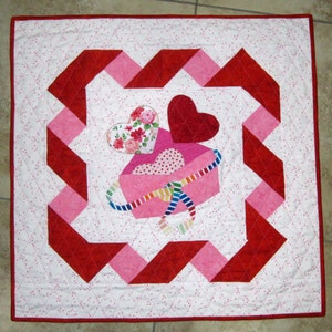 May include: A pink and red quilted heart-shaped box with a red bow and hearts on a white background with pink and red polka dots.