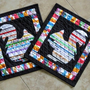 May include: Two black fabric squares with colorful borders and a white silhouette of a squirrel in the center of each square.