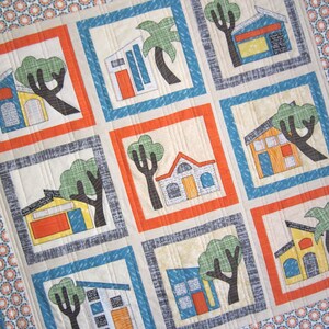 May include: A colorful quilt with a patchwork design featuring houses, trees, and palm trees. The quilt is made of various fabrics in shades of orange, blue, gray, and white.