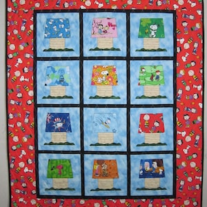 May include: A rectangular quilt featuring a red border patterned with various Peanuts characters. The center contains twelve square blocks, each displaying a lamp with a unique patterned shade and a tan base, set against a light blue background.