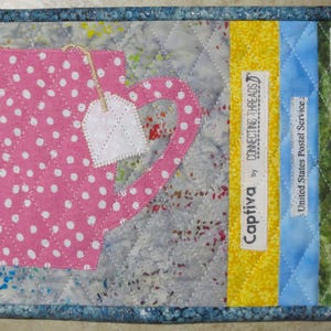 May include: A pink and white polka dot teacup with a white tea bag hanging from the handle. The teacup is on a gray and white background with a yellow, blue, and green striped border. The border has labels that read "Connecting Threads by Captiva", "United States Postal Service", and "Nature's Nursery".