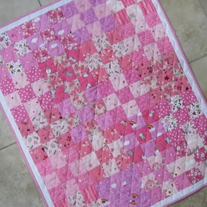 May include: A pink and white patchwork baby quilt with a diamond pattern. The quilt is made of various pink floral fabrics and has a white border.