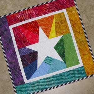 May include: A colorful patchwork quilt with a white star in the center. The quilt is made of different fabrics in shades of red, orange, yellow, green, blue, and purple. The quilt is edged with a black and white striped fabric.
