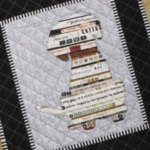 May include: A quilted fabric wall hanging with a black and white striped border. The center of the hanging features a silhouette of a dog made from fabric scraps with text that reads "HI FASHION FABRICS, INC. SPRING 2014", "COPYRIGHT PROTECTED", and "SAUNYITandover created by Kim Schaefer for Andover".