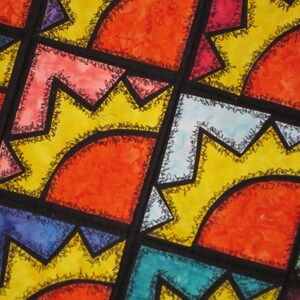 May include: A colorful quilt with geometric shapes in shades of red, orange, yellow, blue, and green. The shapes are outlined in black and have a decorative stitching detail.
