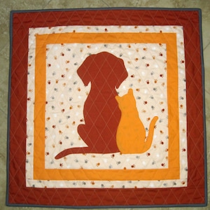 May include: A quilted wall hanging with a brown dog and orange cat silhouette on a cream background with brown paw prints. The background is surrounded by orange and red borders.