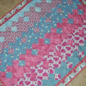 May include: A quilt with a diagonal pattern of pink, blue, and white fabric squares. The quilt has a border of pink and white fabric with a heart design.