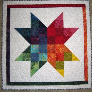 May include: A colorful patchwork quilt with a star design in the center. The star is made up of different colored squares and is surrounded by a white background with small white dots. The quilt is bordered by a dark green, red, and black trim.