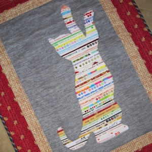 May include: A gray fabric wall hanging with a colorful cat-shaped applique. The applique is made of strips of fabric with various patterns and text, including "Monster Fibers" and "Copyright Protected".