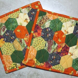May include: Two orange and green quilted pot holders with a fall theme. The fabric features a variety of patterns including corn, leaves, and pumpkins.