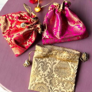 May include: Three small drawstring bags with gold embroidery. The bags are pink, red, and gold. The bags are on a pink surface with string lights and roses.