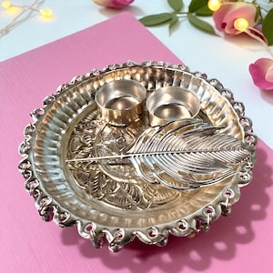 May include: Silver-toned decorative tray with two small candle holders and a feather design. The tray has a scalloped edge and floral detailing. The background is a pink surface with decorative flowers.