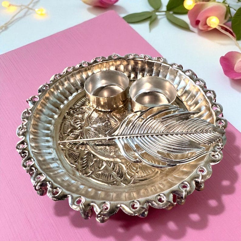 May include: Silver-toned decorative tray with two small candle holders and a feather design. The tray has a scalloped edge and floral detailing. The background is a pink surface with decorative flowers.
