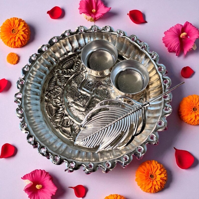 May include: A silver-colored decorative tray with two small cups and a detailed leaf design. The tray is surrounded by orange flowers, pink hibiscus flowers, and red rose petals on a light purple surface.