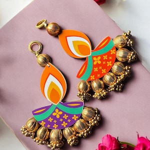 May include: A pair of decorative earrings featuring a colorful diya lamp design. The earrings have an orange, white, and yellow flame, with a purple and green base adorned with gold-colored beads. The earrings are designed with a gold-colored loop for hanging.