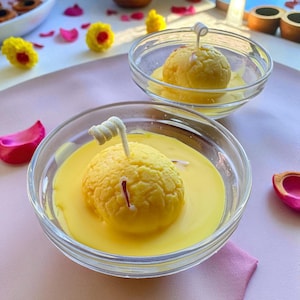 May include: Two clear glass bowls filled with a yellow liquid and a round, yellow dessert. Each dessert has a white, twisted garnish. The bowls sit on a pink surface, with flower petals and small yellow flower decorations scattered around.