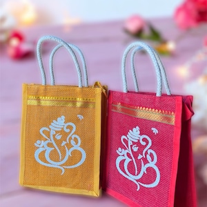 May include: Two small, decorative jute bags with white rope handles. The bags are decorated with a gold trim and a white image of the Hindu deity Ganesha. One bag is yellow and the other is red.