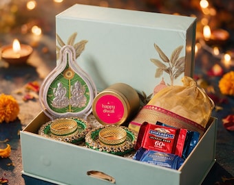 May include: A light green gift box containing a silver-toned Lakshmi and Ganesha idol in a leaf-shaped frame, two green and gold decorative candle holders, a gold tin labeled Happy Diwali, a gold fabric pouch, and Ghirardelli chocolate squares.