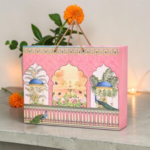May include: A pink gift bag with a decorative design featuring arched windows, peacocks, and floral elements. The bag has a gold rope handle and is set against a neutral background. The bag's design includes a repeating pattern and a detailed border.
