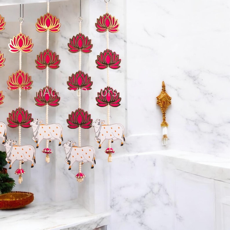 May include: A set of three pink and gold lotus flower and cow door hangings. The hangings are made of paper and are strung together with a white string. The cows are white with black spots and have gold horns. The lotus flowers are pink with gold accents.