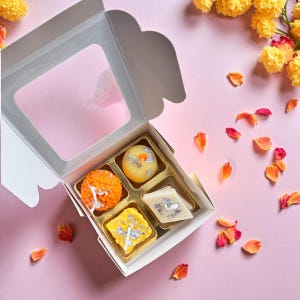 May include: A white box with a clear window, containing four colorful, decorated sweets. The sweets are orange, yellow, and white, with silver leaf accents. The box sits on a pink surface, with scattered flower petals.