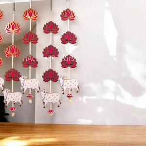 May include: A set of three pink and gold lotus flower garlands with white cows hanging from the bottom. The garlands are strung with white beads and are hanging from the ceiling.