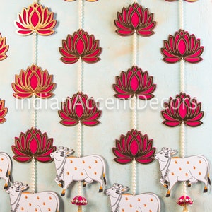 May include: A decorative hanging garland with pink and gold lotus flower cutouts and white cow figures. The garland is strung with white pearl beads and pink tassels.
