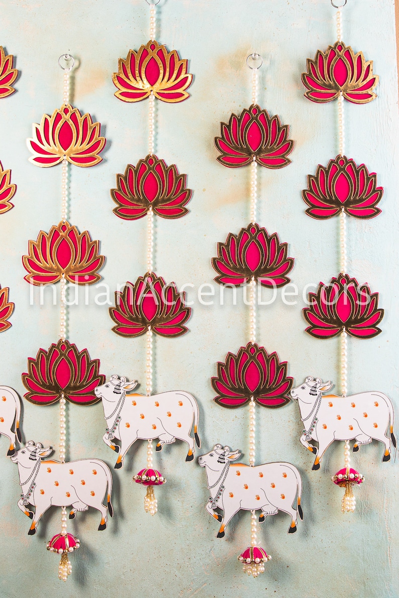 May include: A decorative hanging garland with pink and gold lotus flower cutouts and white cow figures. The garland is strung with white pearl beads and pink tassels.