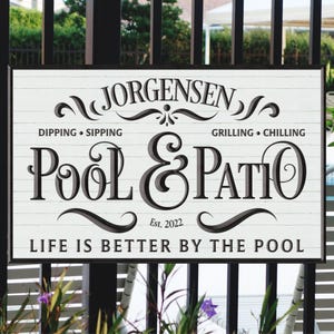 May include: White wooden sign with black text that reads "Jorgensen Pool & Patio Est. 2022 Life is Better by the Pool". The sign has a decorative flourish around the words "Pool & Patio" and the words "Dipping Sipping" and "Grilling Chilling" are written above and below the main text.