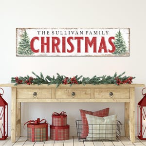 May include: A rectangular wooden sign with the text "THE SULLIVAN FAMILY CHRISTMAS" in red letters. The sign is above a wooden table decorated with a garland, red lanterns, and wrapped gifts. The sign has two green Christmas trees.