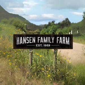 May include: A wooden sign for Hansen Family Farm, established in 1989, stands in a field of tall grass and wildflowers. The sign is dark brown with white lettering and is mounted on wooden posts. The background features a road, trees, and a cloudy sky.